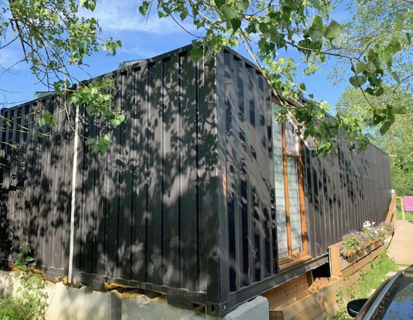 Two grey shipping containers that form a house with wooden windows sit in the shade of a tree.
