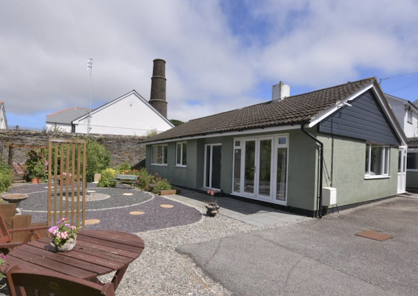 A green detached bungalow is pictured on a large driveway with flower pots and ornamental paving.