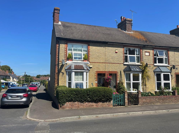 An end of terrace house with red brick detailing and plants is pictured with cars parked in the road to the left of it