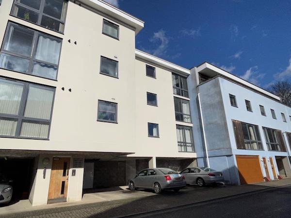 Cars are parked beneath a modern block of flats. There are blue skies overhead