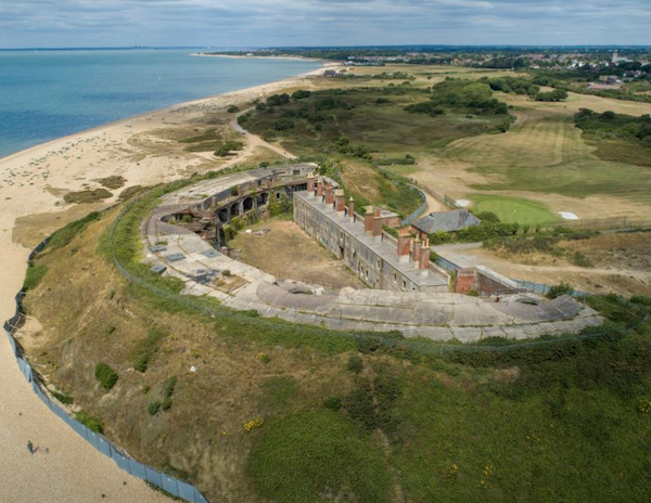 A semicircular victorian fort by the sea is pictured from above with greenery surrounding it and the coastline on the horizon